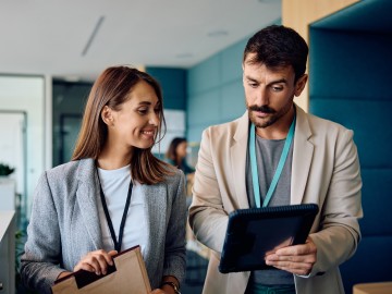 Cyberkriminalität auf dem Vormarsch - Risiken verstehen und absichern: Eine Frau und ein Mann in einem Büro. Er hält ein Tablet und zeigt ihr etwas darauf.