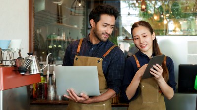 So reagieren Betriebe richtig auf schlechte Bewertungen und Fake-Rezensionen: Zwei Mitarbeiter eines Cafes diskutieren, was sie auf einem Laptop und Tablet sehen.