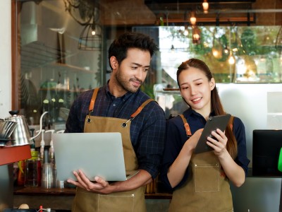 So reagieren Betriebe richtig auf schlechte Bewertungen und Fake-Rezensionen: Zwei Mitarbeiter eines Cafes diskutieren, was sie auf einem Laptop und Tablet sehen.
