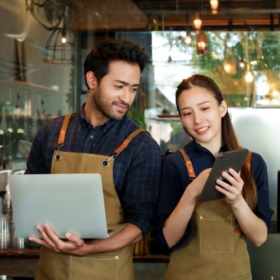 So reagieren Betriebe richtig auf schlechte Bewertungen und Fake-Rezensionen: Zwei Mitarbeiter eines Cafés diskutieren, was sie auf einem Laptop und Tablet sehen.