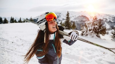 Von Anreise bis Après Ski - Gut abgesichert im Skiurlaub: Frau trägt Ski auf den Schultern und lacht