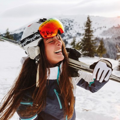 Von Anreise bis Après Ski - Gut abgesichert im Skiurlaub: Frau trägt Ski auf den Schultern und lacht