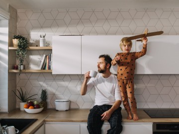 Zurück in Schule, Kita und Alltag - So schützen Sie Kinder und Familie: Vater trinkt in der K&uuml;che Kaffee und der Sohn spielt auf der Arbeitsplatte mit einem Modellflugzeug