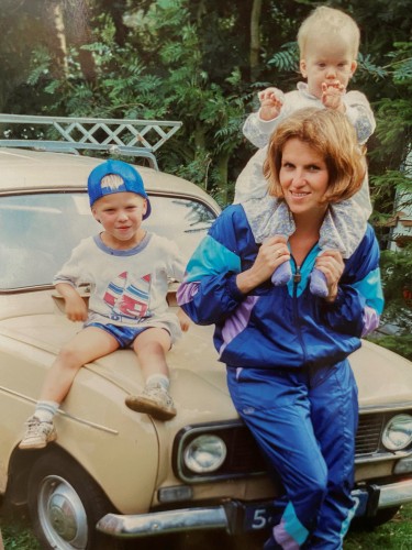 Frau mit zwei kleinen Kindern sitzt auf einem alten Auto im Look der 80er Jahre