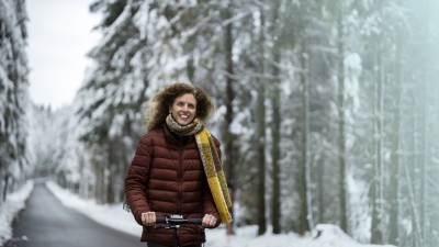 7 Tipps, die Sie beim E-Scooter-Fahren im Winter beachten sollten: Frau in Daunenjacke und Schal f&auml;hrt mit einem E-Scooter durch eine Winterlandschaft