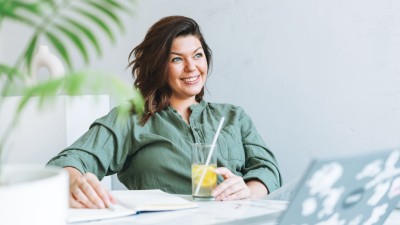 Rente mit 35? Das steckt hinter Frugalismus. : Frau in grüner Bluse führt Haushaltsbuch und trink Zitronenwasser