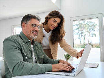 Versicherungen und Finanzen 2026 - Das ändert sich : Ein l&auml;chelnder Mann mittleren Alters arbeitet am Computer, w&auml;hrend seine Frau neben ihm  steht.