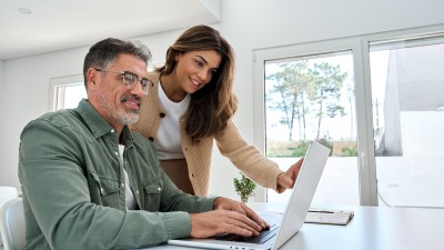 Versicherungen und Finanzen 2026 - Das ändert sich : Ein l&auml;chelnder Mann mittleren Alters arbeitet am Computer, w&auml;hrend seine Frau neben ihm  steht.