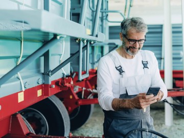 Ein Landwirt sieht auf sein Smartphone, im Hintergrund stehen landwirtschaftliche Anhänger