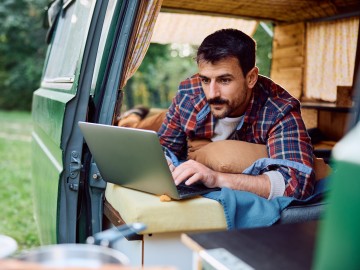 Van-Ausbau - Wohnmobilversicherung für den Campervan?: Mann schaut beim Camping aus dem Camping-Bus auf seinen Laptop