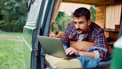 Van-Ausbau - Wohnmobilversicherung für den Campervan?: Mann schaut beim Camping aus dem Camping-Bus auf seinen Laptop