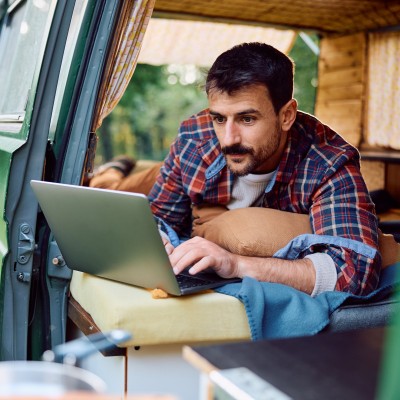 Van-Ausbau - Wohnmobilversicherung für den Campervan?: Mann schaut beim Camping aus dem Camping-Bus auf seinen Laptop