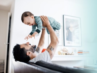 Mann sitzt auf der Couch und hält ein lachendes Baby hoch.