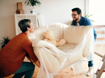 Private Haftpflichtversicherung: Eine Frau und ein Mann schleppen beim Umzug einen wei&szlig;en Sessel in Folie auf dem ein Teddy sitzt. 