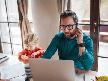 Rechtsschutzversicherung: Mann telefoniert mit Laptop vor sich und Kleinkind isst seinen Brei aus einem Schüsselchen. 