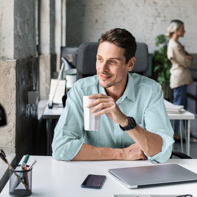 Mann sitzt im B&uuml;ro an seinem Schreibtisch mit einem Trinkbecher in der Hand