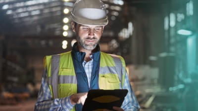 Mitarbeiter mit Schutzhelm im Betrieb schaut auf Tablet