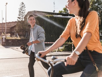 Ein Frau auf einem Fahrrad und ein Mann mit Skateboard lächeln sich an