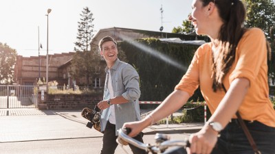 Ein Frau auf einem Fahrrad und ein Mann mit Skateboard lächeln sich an
