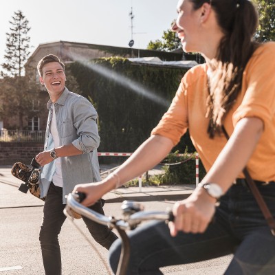 Ein Frau auf einem Fahrrad und ein Mann mit Skateboard lächeln sich an