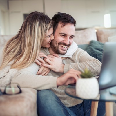 Pflegefall - So schützen Sie Ihr Haus & Vermögen: Junges Paar sitzt lachend und eng umschlungen vor einem Laptop