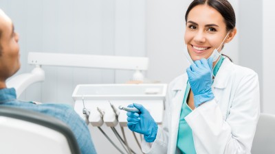 Eine Zahn&auml;rztin mit Bohrer in der Hand zieht ihren Mundschutz herunter und l&auml;chelt