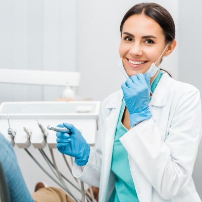 Eine Zahnärztin mit Bohrer in der Hand zieht ihren Mundschutz herunter und lächelt