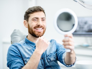 Ein Patient sitzt im Zahnarztstuhl und betrachtet seine Z&auml;hne in einem Handspiegel