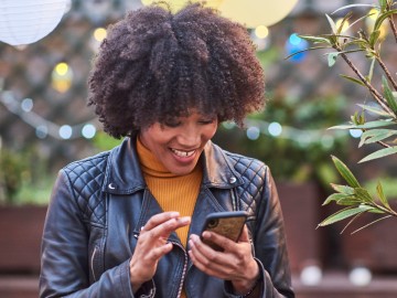 Frau mit Afrolook und mit Lederjacke bedient lächelnd Ihr Smartphone