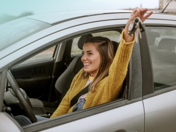 Kfz-Familienfahrer-Versicherung: Junge Frau sitzt glücklich in einem Auto und streckt einen Arm aus dem Fenster