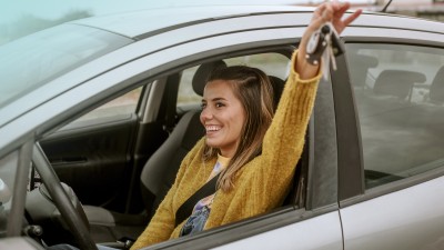 Kfz-Familienfahrer-Versicherung: Junge Frau sitzt glücklich in einem Auto und streckt einen Arm aus dem Fenster