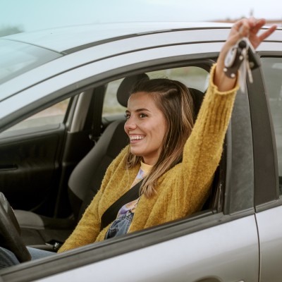 Kfz-Familienfahrer-Versicherung: Junge Frau sitzt glücklich in einem Auto und streckt einen Arm aus dem Fenster