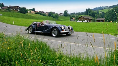 Ein dunkelblauer Morgan-Oldtimer fährt durch eine alpine Dorflandschaft.