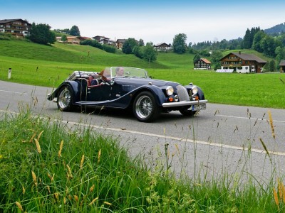 Oldtimer-Saisonkennzeichen: Ein dunkelblauer Morgan-Oldtimer fährt durch eine alpine Dorflandschaft.