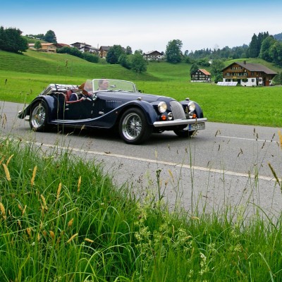 Ein dunkelblauer Morgan-Oldtimer fährt durch eine alpine Dorflandschaft.