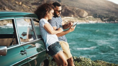 Paar lehnt am Auto auf einer Klippe am Meer und schaut aufs Handy