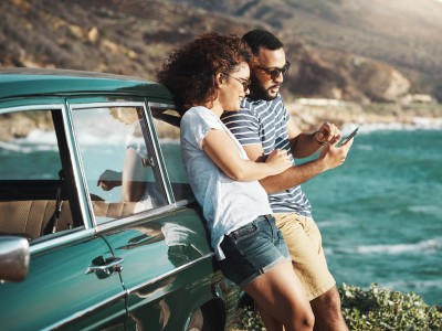 Paar lehnt am Auto auf einer Klippe am Meer und schaut aufs Handy