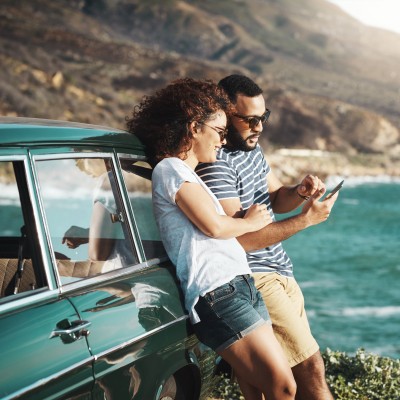 Paar lehnt am Auto auf einer Klippe am Meer und schaut aufs Handy