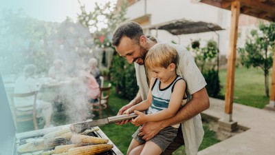 Wohngebäudeversicherung: Mann steht vor einem Grill und hält ein kleines Kind auf dem Arm