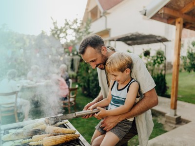 Wohngebäudeversicherung: Mann steht vor einem Grill und hält ein kleines Kind auf dem Arm