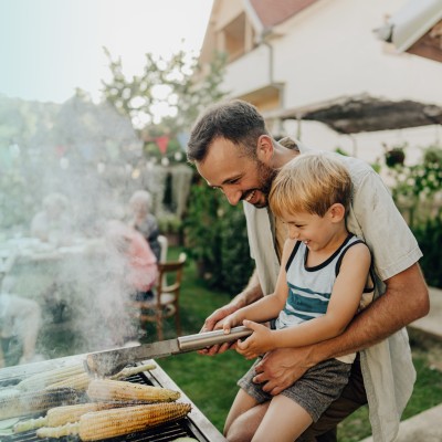 Wohngebäudeversicherung: Mann steht vor einem Grill und hält ein kleines Kind auf dem Arm