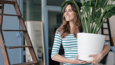 Mietkautionsversicherung: Junge Frau steht in einer Wohnung und hält einen Blumentopf mit einer Pflanze