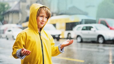 Elementarversicherung: Kind im Regenmantel steht in starkem Regen auf Straße