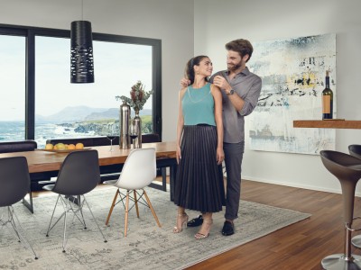 Hausratversicherung für Paare & Partner: Mann legt im Esszimmer Frau Halskette an, im Hintergrund, Küstenlandschaft