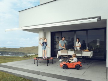 Hausratversicherung für Familien: Kinder spielen auf Terasse während Eltern den Tisch decken, im Hintergrund Küstenlandschaft