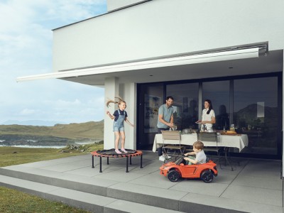 Hausratversicherung für Familien: Kinder spielen auf Terasse während Eltern den Tisch decken, im Hintergrund Küstenlandschaft