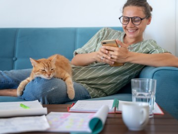 Eine Frau sitzt gemütlich mit Ihrer Katze und einem Smartphone auf dem Sofa 