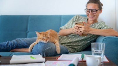Eine Frau sitzt gemütlich mit Ihrer Katze und einem Smartphone auf dem Sofa 