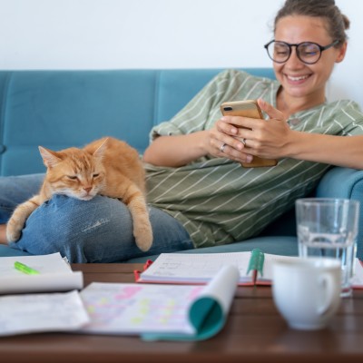 Eine Frau sitzt gemütlich mit Ihrer Katze und einem Smartphone auf dem Sofa 