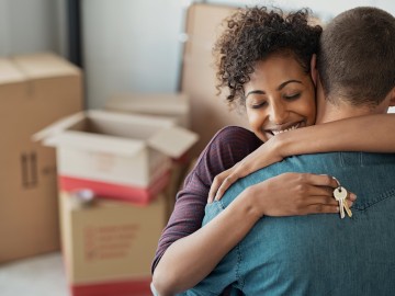 Junges Paar umarmt sich mit Schlüssel in der Hand. Im Hintergrund Umzugskartons. 